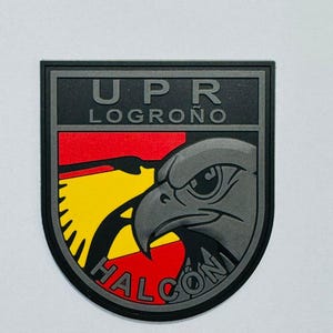 May include: A black and gray patch with the text "UPR LOGRO&Ntilde;O" at the top. The patch features a stylized gray eagle head over a red, yellow, and black background. The word "HALCON" is written at the bottom.