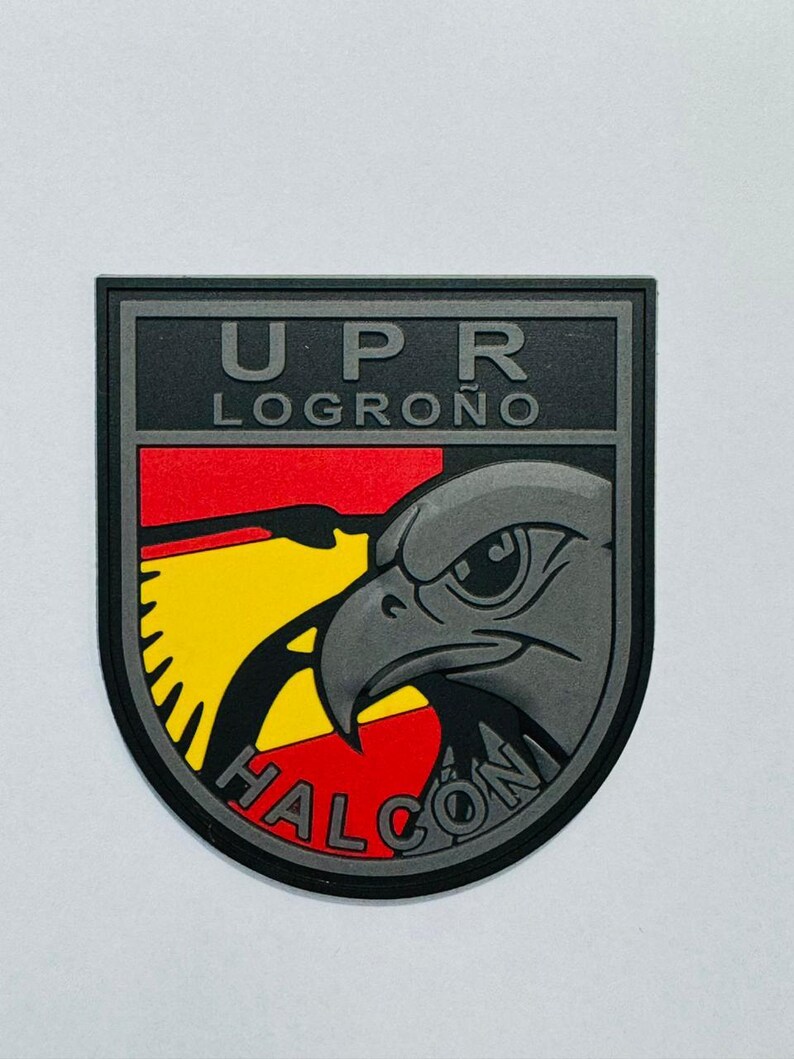 May include: A black and gray patch with the text "UPR LOGRO&Ntilde;O" at the top. The patch features a stylized gray eagle head over a red, yellow, and black background. The word "HALCON" is written at the bottom.