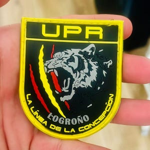 May include: A yellow and black patch with the text "UPR" at the top. The patch features a roaring tiger graphic and the words "LA LINEA DE LA CONCEPCION" and "LOGRO&Ntilde;O". Red and yellow claw marks are on the left side.