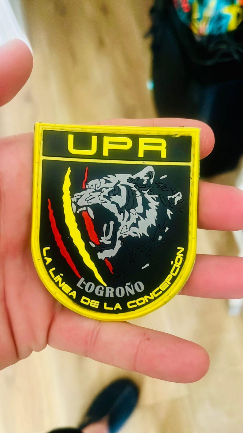 May include: A yellow and black patch with the text "UPR" at the top. The patch features a roaring tiger graphic and the words "LA LINEA DE LA CONCEPCION" and "LOGRO&Ntilde;O". Red and yellow claw marks are on the left side.