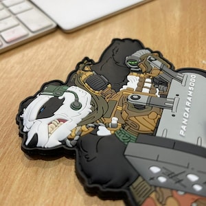 Pu&ograve; includere: Una toppa dettagliata raffigurante un personaggio a fumetti simile a un tasso in tenuta militare, con un'arma di grandi dimensioni. La toppa &egrave; prevalentemente nera, con accenti di bianco, marrone e verde. Il testo "PANDARAM5000" &egrave; visibile.