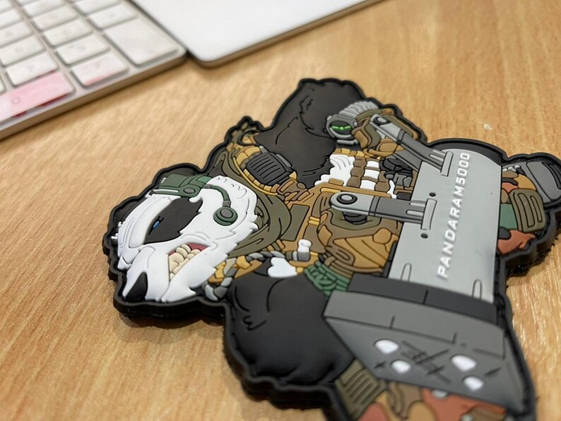 Pu&ograve; includere: Una toppa dettagliata raffigurante un personaggio a fumetti simile a un tasso in tenuta militare, con un'arma di grandi dimensioni. La toppa &egrave; prevalentemente nera, con accenti di bianco, marrone e verde. Il testo "PANDARAM5000" &egrave; visibile.