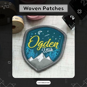 Könnte beinhalten: Ein schildförmiger gewebter Aufnäher mit dem Text "Ogden Utah" in gelber Schrift. Der Aufnäher hat einen teal-farbenen Hintergrund mit einem Berg- und Baumdesign. Der Aufnäher ist mit grauem Faden eingefasst.