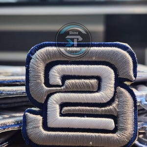 May include: Embroidered patch with the number "2" in a stylized design. The patch has a white centre, surrounded by grey and outlined in navy blue. The patch has the text "Shine Patches" in a circular logo.