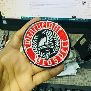 May include: A round, embroidered patch with a red border and black and white center design. The patch features the text "TWENTY EIGHT BROTHERS" in a circular pattern around a central emblem. The emblem includes a helmet and laurel leaves.