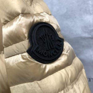 Op de afbeelding: Close-up van een goudkleurige gewatteerde jas mouw. Een zwarte cirkelvormige patch met de merknaam is op de mouw genaaid. De jas heeft een glanzende, gladde textuur.