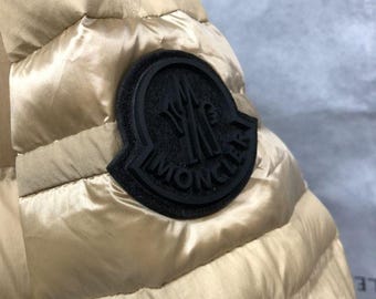 May include: Close-up of a gold-colored quilted jacket sleeve. A black circular patch with the brand name is sewn onto the sleeve. The jacket has a shiny, smooth texture.