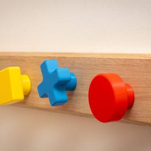Può includere: Appendiabiti da parete in legno con quattro pomelli colorati. I pomelli hanno la forma di un triangolo verde, un quadrato giallo, una croce blu e un cerchio rosso. Il supporto è in legno chiaro.