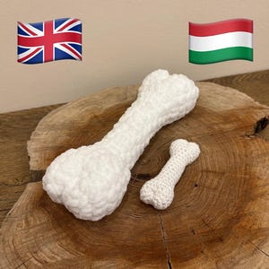 Puede incluir: Dos huesos de perro blancos, tejidos a ganchillo, de diferentes tamaños, descansan sobre una superficie de madera. El hueso más grande tiene un extremo bulboso, mientras que el más pequeño es una versión en miniatura. La textura del hilo es visible.