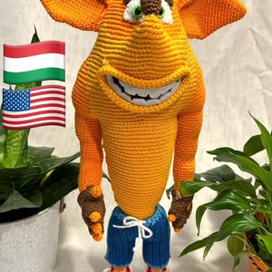 Puede incluir: Figura de ganchillo hecha a mano de un personaje de dibujos animados en tonos naranja, marrón y blanco. La figura tiene orejas grandes, una amplia sonrisa y lleva pantalones cortos azules y zapatillas rojas. La figura mide unos 30 cm de alto.