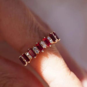 Pode incluir: Um anel de ouro com um padrão alternado de rubis vermelhos retangulares em lapidação baguette e pares de pequenos diamantes brancos redondos em lapidação brilhante. As pedras estão cravadas numa fila contínua no topo do aro.