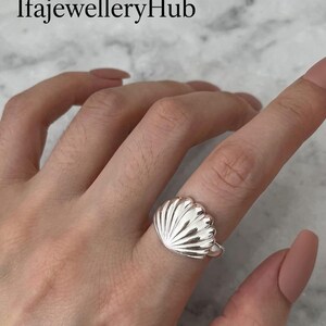 Peut inclure: Une bague en argent poli présentant un motif de coquillage proéminent avec des crêtes verticales. La bague est portée sur un doigt et le texte IfajewelleryHub est visible dans le coin supérieur gauche. Le métal a une surface réfléchissante.