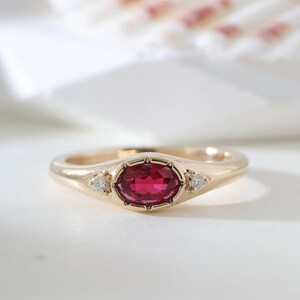 Può includere: Un anello in oro con una pietra preziosa centrale di colore rosa intenso dal taglio ovale, incastonata con piccole griffe. La fascia include due minuscole pietre bianche rotonde, una su ciascun lato dell'incastonatura centrale, integrate nell'anello.