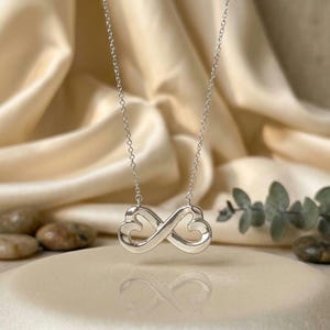 Puede incluir: Un collar de tono plateado con un colgante en forma de símbolo de infinito compuesto por dos contornos de corazón interconectados. El colgante cuelga de una cadena fina y delicada de tono plateado, con un acabado metálico pulido y brillante.