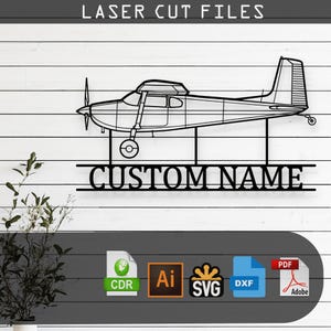 May include: Black metal laser-cut airplane wall art with the text "LASER CUT FILES" at the top. Below the plane is the text "CUSTOM NAME". The airplane has a propeller, wings, and tail. The background is white horizontal planks.