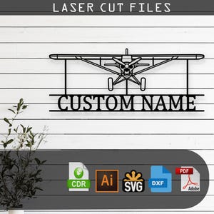 Op de afbeelding: Zwart metalen lasergesneden bord met een vliegtuigontwerp en de tekst "CUSTOM NAME" eronder. De woorden "LASER CUT FILES" staan bovenaan. Het bord staat tegen een witte houten achtergrond.