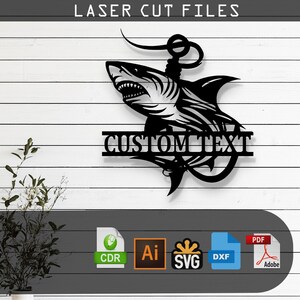 May include: Black metal laser-cut wall art featuring a shark design with an anchor and the words "CUSTOM TEXT". The artwork is mounted on a white wooden plank background. The top of the image reads "LASER CUT FILES".