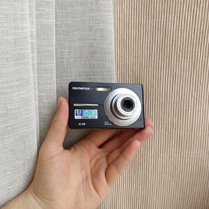 May include: A black Olympus X-15 digital camera with a silver lens barrel. The front of the camera features a flash, a lens, and a small rectangular label with the text 8.0 Megapixels and the Olympus logo printed in white on the top left corner.