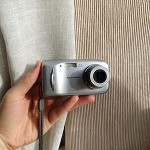 Op de afbeelding: Zilveren Olympus digitale camera met een zwarte lens en de tekst "OLYMPUS". De camera heeft een ingebouwde flitser en een polsband. Het is een 4.0 megapixel model.