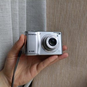 Puede incluir: Una cámara digital plateada Olympus X-43 con un objetivo de zoom óptico de 5x, un flash integrado y una correa de muñeca gris. La parte frontal del cuerpo de la cámara muestra la marca Olympus y el modelo X-43 en letras negras.