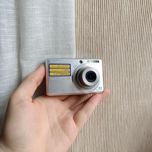 Puede incluir: Una cámara digital Sony Cyber-shot plateada con una lente y un flash prominentes. La cámara tiene una pequeña pantalla y varios botones. La cámara se sostiene en una mano.