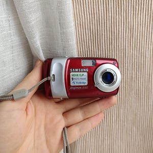 May include: A red and silver Samsung Digimax A502 digital camera. The camera has a lens, flash, and a wrist strap. The camera has the words "Movie Clip", "Photo Frame", and "PictBridge" printed on it.