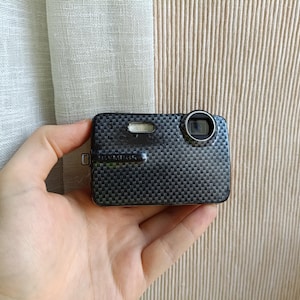 Puede incluir: Una cámara digital compacta con un patrón de fibra de carbono gris oscuro texturizado. La cámara tiene una lente, un flash y la marca "OLYMPUS" es visible. La cámara se sostiene en una mano.