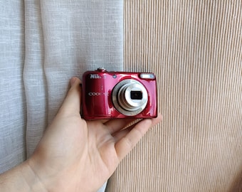 May include: A red Nikon Coolpix digital camera. The camera has a silver lens and a built-in flash. The camera is held in a hand against a neutral background.