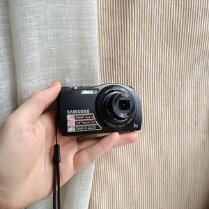 May include: A black Samsung digital camera with a 5x zoom lens is held in a hand. The camera has a built-in flash and a 3.0-inch touch LCD screen. The camera's strap is black.