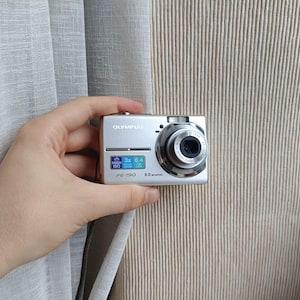 May include: A silver Olympus FE-190 digital camera with a 6.0 megapixel sensor. The front of the camera features a lens, a flash, and a small display area with text reading High ISO, 3x Optical Zoom, and 6.4. The model name FE-190 is printed below.