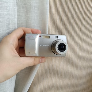May include: A silver Olympus C-310 Zoom digital camera featuring a 3.2 megapixel sensor. The front of the device displays the brand name Olympus, the model number, and a lens with the text Olympus Lens AF Zoom 5.8-17.4mm 1:2.8-4.8.