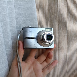 May include: A silver Kodak digital camera featuring a retractable lens with text reading Kodak AF 3x Optical Aspheric Lens 36mm-108mm Equivalent. The compact device includes a built-in flash, a metallic finish, and a thin gray wrist strap attached to the side.