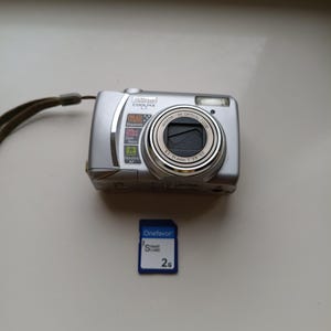 May include: A silver Nikon Coolpix L1 digital camera with a 5x optical zoom Nikkor lens, 6.2 megapixels, and a wrist strap, accompanied by a blue Onefavor 2G Smart Card memory card with white text and a small logo.