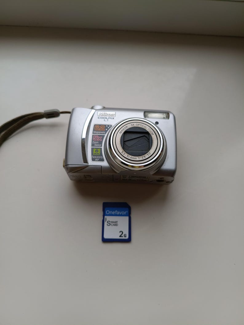 May include: A silver Nikon Coolpix L1 digital camera with a 5x optical zoom Nikkor lens, 6.2 megapixels, and a wrist strap, accompanied by a blue Onefavor 2G Smart Card memory card with white text and a small logo.