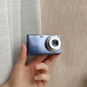 May include: A blue Nikon Coolpix digital camera with a brushed metal finish. The front of the camera features the Nikon logo, the Coolpix brand name, a built-in flash, and a circular lens barrel with text indicating a 5x optical zoom capability.