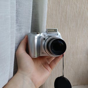 May include: A silver digital camera featuring a prominent lens with 10x optical zoom capability. The camera body includes a pop-up flash unit on the top, a shutter button, and a lens cap attached by a thin black cord hanging below the device.
