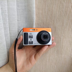 Puede incluir: Una cámara digital Pentax Optio H90 con cuerpo plateado y naranja. La parte frontal presenta un objetivo negro, un flash y texto que indica 12,1 megapíxeles, zoom óptico de 5x, vídeo HD y detección de rostros. Una correa de muñeca negra está sujeta al lateral.