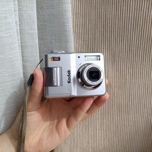 May include: A silver Kodak digital camera featuring a rectangular body, a central lens with text reading Kodak AF 3x Optical Aspheric Lens, a built-in flash, and a small Kodak Color Science logo on the top left corner, accompanied by a wrist strap.
