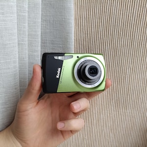 May include: A compact digital camera with a metallic lime green body and a black side panel. The front features a silver lens ring, a built-in flash, and the text Kodak 12 Megapixels printed in white on the green surface near the lens.