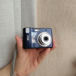 Op de afbeelding: Een donkerblauwe Nikon Coolpix digitale camera met een zilverkleurige lenscilinder. De voorzijde bevat een kleine sticker met de tekst PC AR 46 en een zilverkleurig label met de tekst 1000, 1000, 2.4 en 7.1 Megapixels. Een grijze polsband is bevestigd.