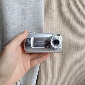 以下が含まれることがあります： 7.1メガピクセルのセンサーと3.4倍光学ズームレンズを搭載したシルバーのキヤノンPowerShot A470デジタルカメラ。デバイスの前面には、ブランド名、モデル番号、およびレンズの技術仕様が黒いテキストで印刷されています。