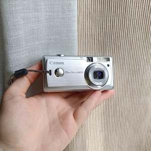 以下が含まれることがあります： リストストラップ付きのシルバーCanon PowerShot A400デジタルカメラ。カメラにはレンズ、フラッシュ、Canonのロゴがあります。カメラは手で持たれています。