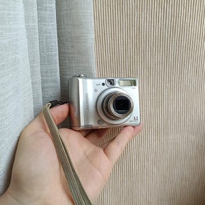 May include: A silver Canon PowerShot A510 digital camera featuring a 3.2 megapixel lens, a built-in flash, and a beige wrist strap. The front of the camera displays the brand name, model, and technical specifications printed in black text.