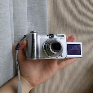 May include: A silver Canon PowerShot A610 digital camera featuring a 5.0 megapixel sensor, a retractable zoom lens with 4x optical zoom, a built-in flash, and a side-hinged LCD screen. A grey wrist strap is attached to the side of the camera body.