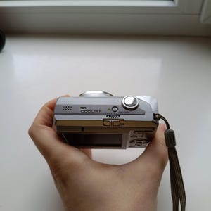 May include: A silver Nikon Coolpix digital camera featuring a top-mounted power button, a speaker grille, and a mode selection switch. The device includes a wrist strap attached to the side and a small green indicator light near the power button.