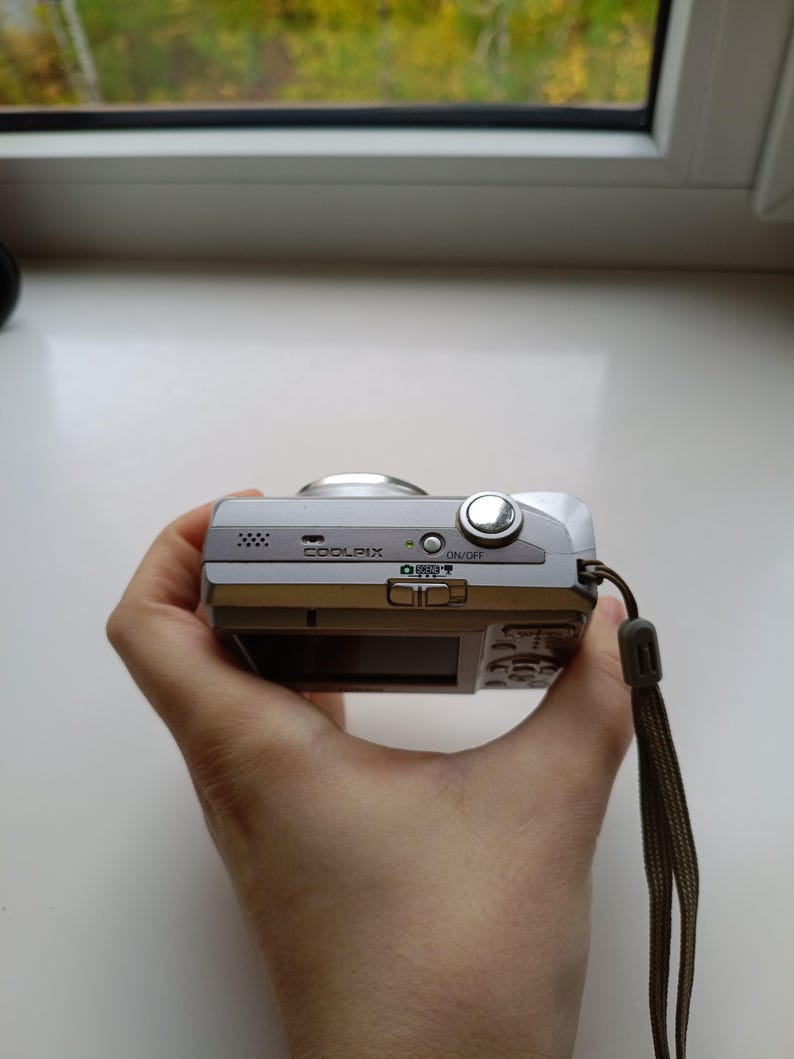 May include: A silver Nikon Coolpix digital camera featuring a top-mounted power button, a speaker grille, and a mode selection switch. The device includes a wrist strap attached to the side and a small green indicator light near the power button.