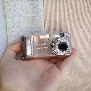 May include: A silver Canon PowerShot A460 digital camera with a 4x zoom lens. The camera has a built-in flash and a 5.0 MP resolution. The camera is held in a hand.