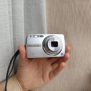 Puede incluir: Una cámara digital Olympus de color plateado con lente retráctil, flash integrado y correa de muñeca negra. La parte frontal muestra la marca Olympus, el nombre del modelo y las especificaciones técnicas de la lente impresas alrededor.