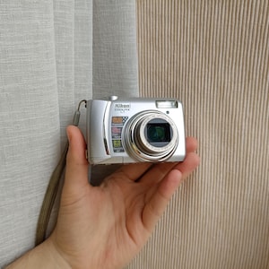 May include: A silver Nikon Coolpix L1 digital camera featuring a 5x optical zoom lens, a built-in flash, and a wrist strap. The front of the camera displays the Nikon Coolpix L1 logo along with labels for 5x optical zoom and other features.