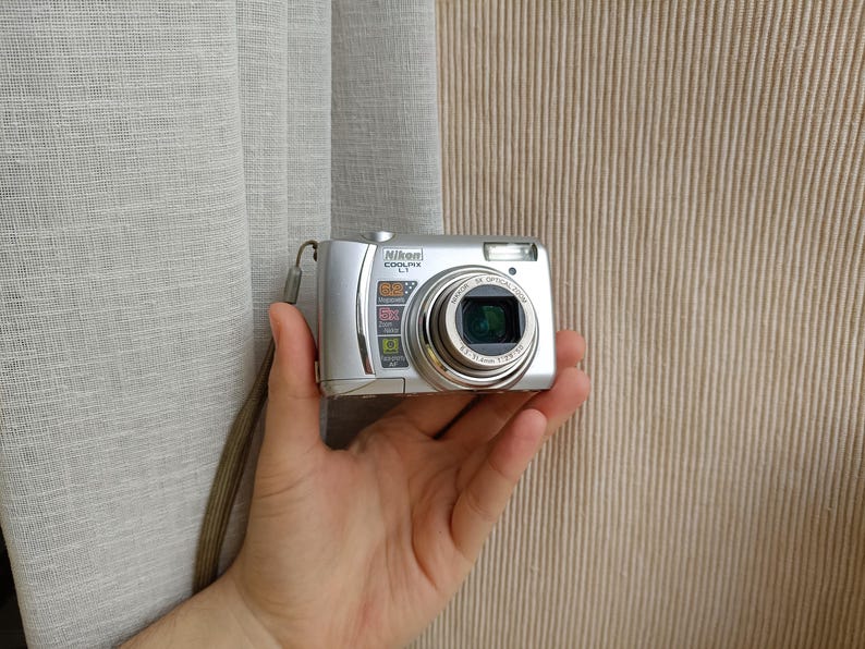 May include: A silver Nikon Coolpix L1 digital camera featuring a 5x optical zoom lens, a built-in flash, and a wrist strap. The front of the camera displays the Nikon Coolpix L1 logo along with labels for 5x optical zoom and other features.